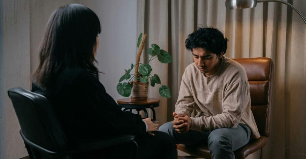 A man sitting in a brown chair, looking down with clasped hands, across from a woman in a black shirt who is partially obscured, in a therapy session.