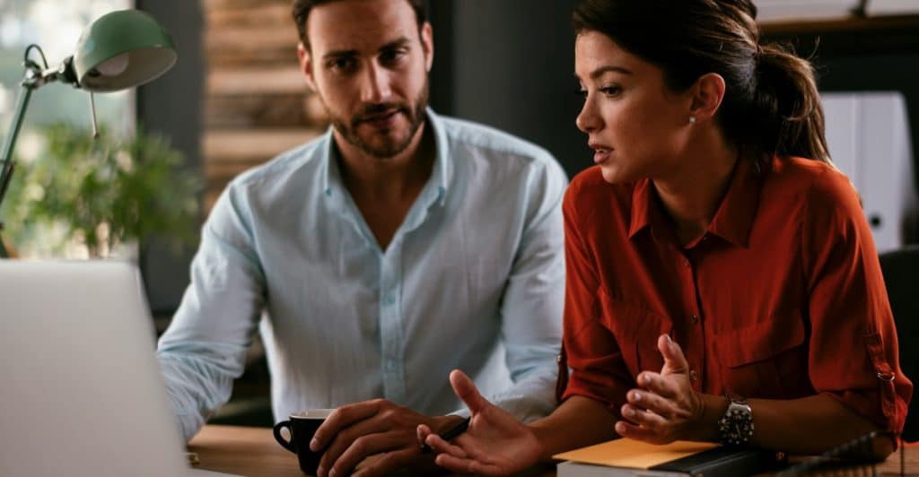A man and a woman sitting at a desk, looking at a laptop and discussing something intently