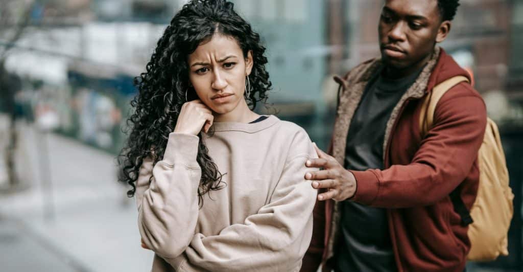 A woman with curly hair looking upset, standing outdoors while a man behind her places a hand on her shoulder.