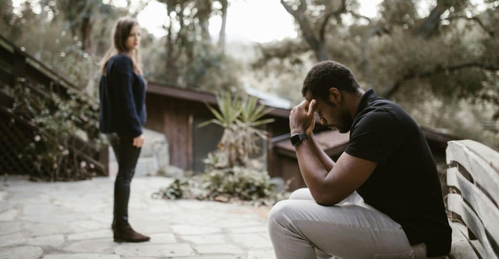 A man sitting on a bench with his head in his hands, with a woman standing in the background.