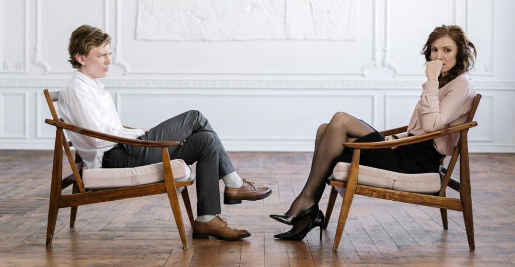 A man and a woman sitting on chairs facing each other in a room, with the woman looking away and the man looking at her.
