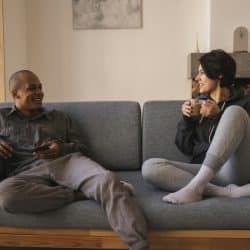 A man and a woman are sitting on a gray couch, facing each other and talking. The man is on the left, holding a remote, and the woman is on the right, holding a mug.