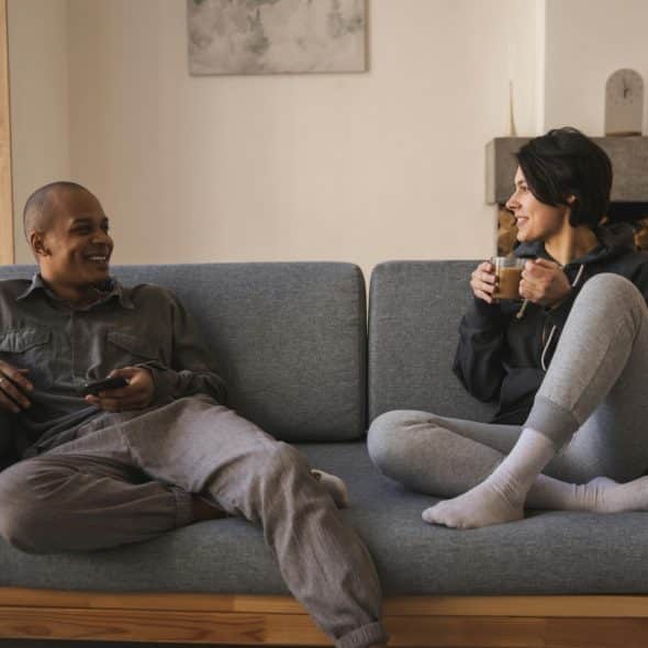 A man and a woman are sitting on a gray couch, facing each other and talking. The man is on the left, holding a remote, and the woman is on the right, holding a mug.