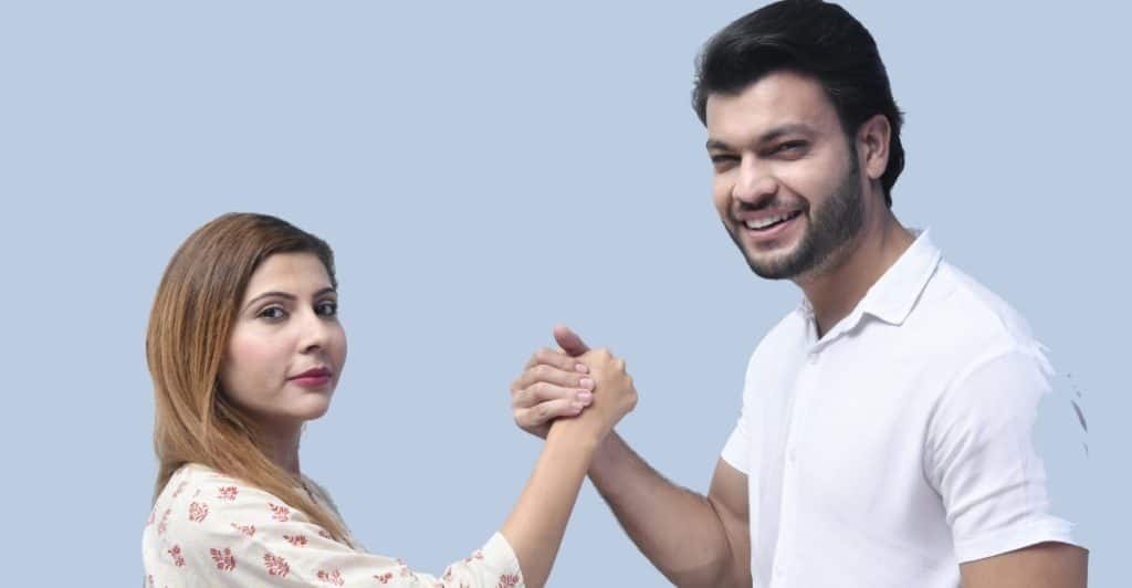 A man and a woman are standing and shaking hands, with the man smiling at the camera.