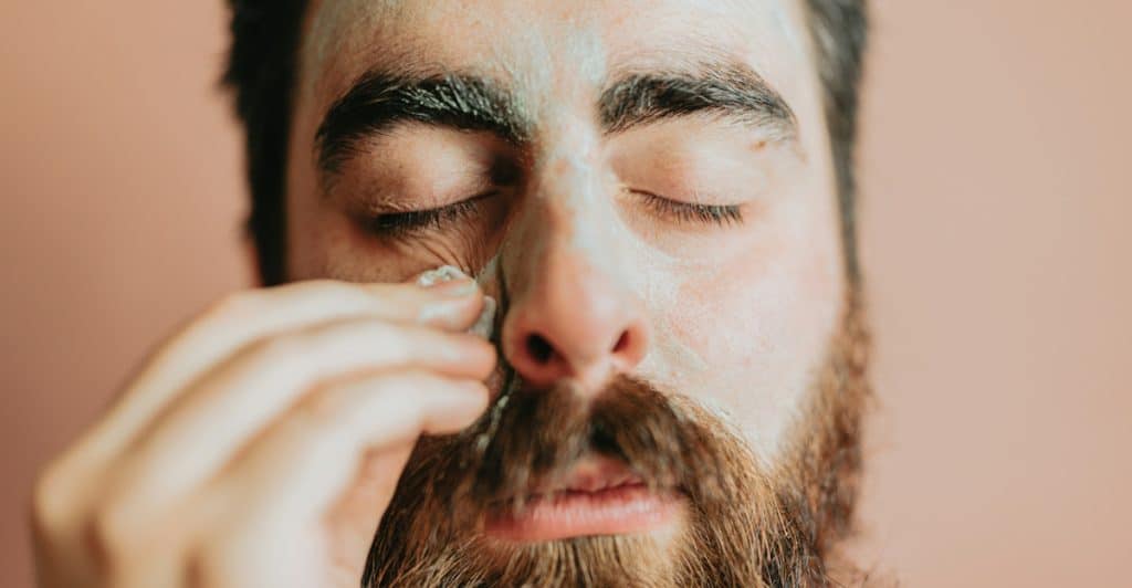 A close-up of a bearded man with his eyes closed applying a cosmetic product to his face.