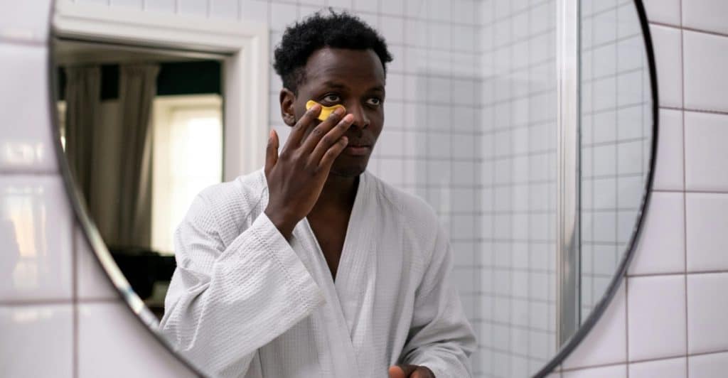 A man applying eye cream under his eye while looking in a mirror.