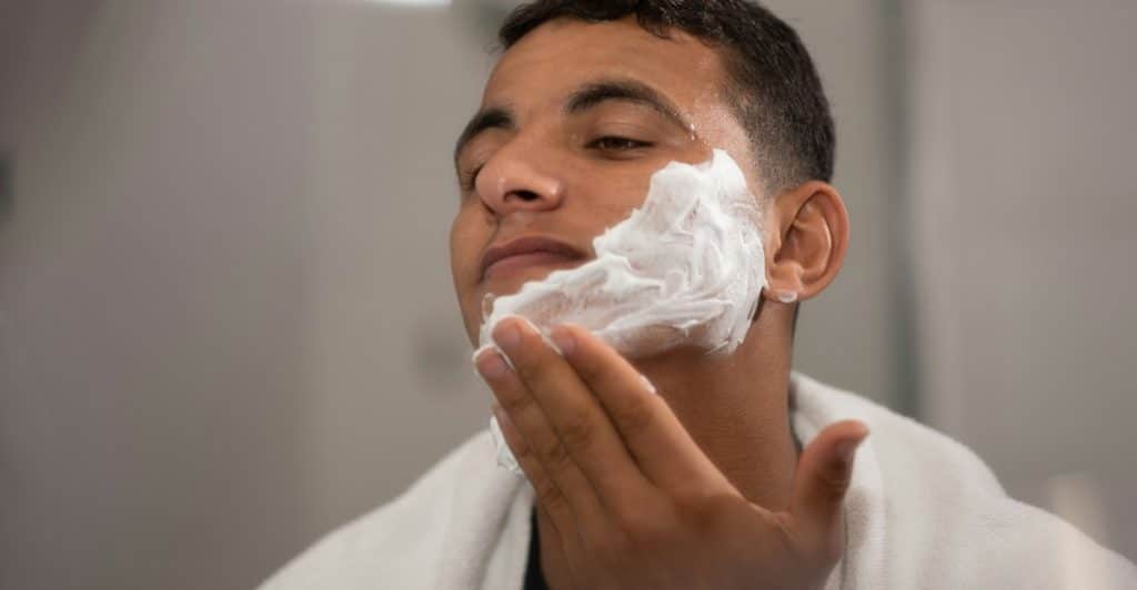 A young man with a towel around his neck applying white shaving foam to his face.