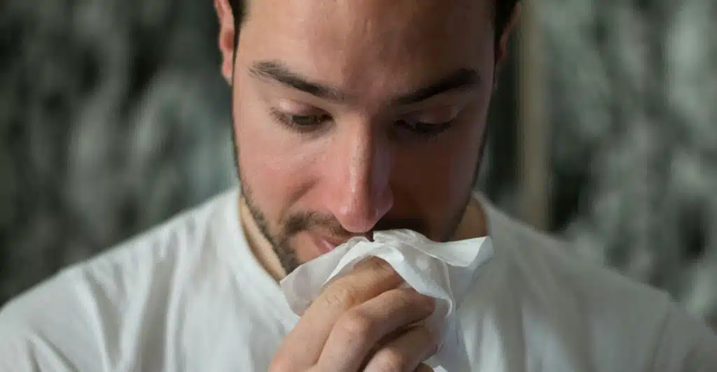 A man wearing a whit t-shirt blotting his face with a white tissue.