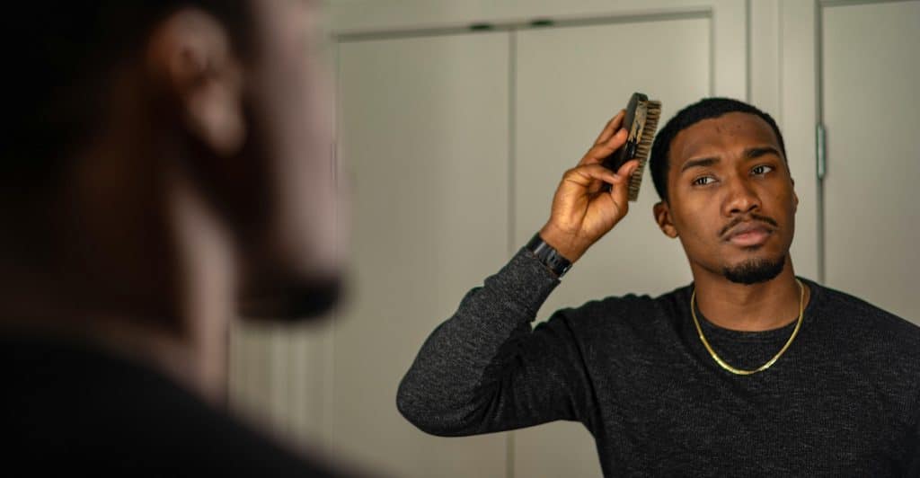 A man with short dark hair and a beard, wearing a dark long-sleeved shirt and a gold chain, brushing his hair in front of a mirror.