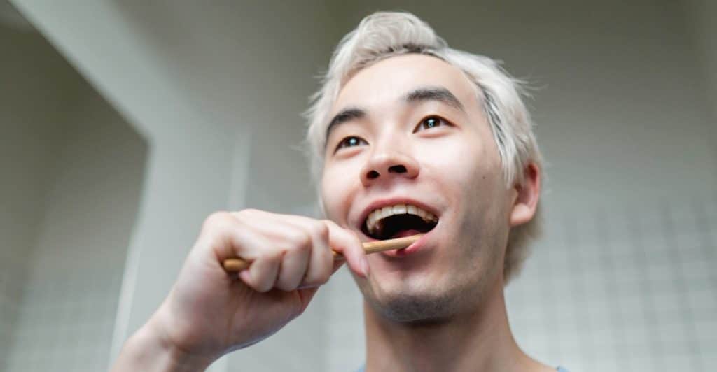 A man with dyed hair brushing his teeth with a wooden toothbrush and smiling.