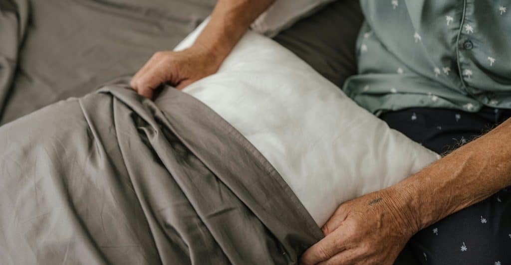 A person's hands putting a white pillow into a gray pillowcase on a bed with gray sheets.