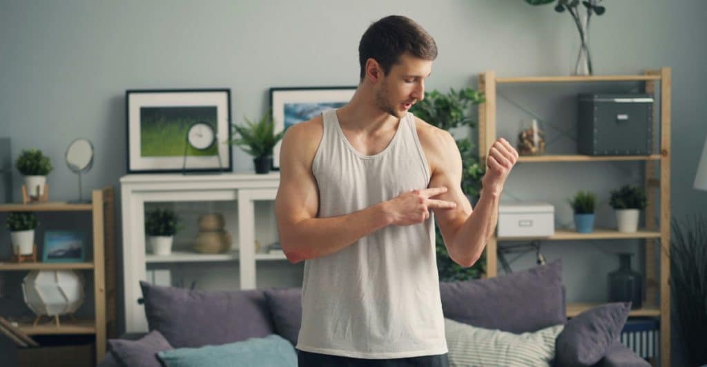 A young man in a white tank top standing in a living room, smiling slightly and pointing at his flexed bicep with his left hand.