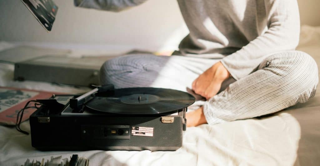 A person sitting on a bed, placing a vinyl record on a turntable with other records nearby.