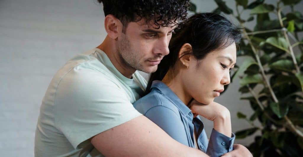 A man embracing a woman from behind, with both looking somber or sad.