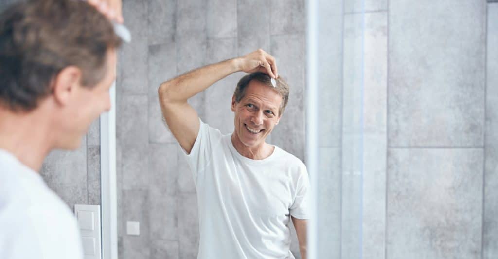 A man in a white t-shirt is looking at his reflection in a bathroom mirror, smiling and touching his hair.