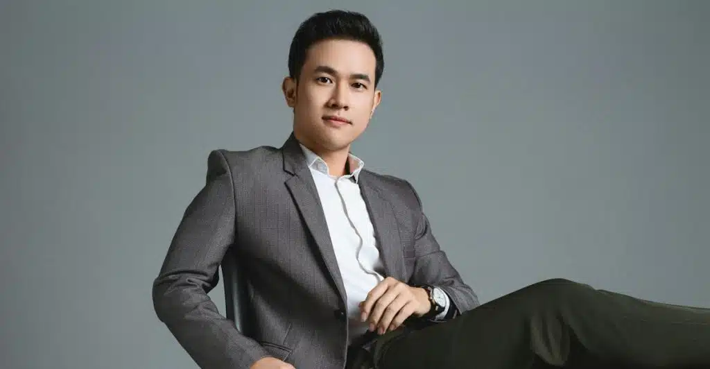 A man sitting in a chair and confidently posing in a grey suit and dark pants against a plain background.