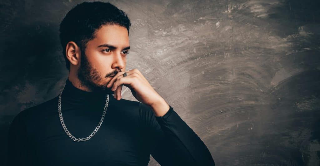 A man in a black turtleneck and silver chain, with a beard and mustache, looking to the side with his hand on his chin.