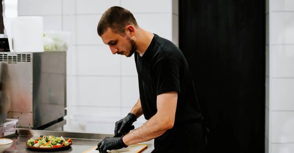 A man in a black t-shirt and black gloves is chopping food on a counter in a kitchen.