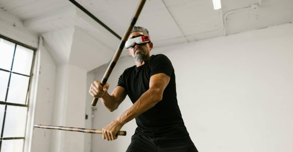 A man with a beard wearing a VR headset and a black t-shirt and pants, holding two sticks in a fighting pose in a white room with large windows.