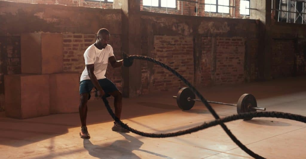 A man in a white t-shirt and blue shorts is vigorously swinging heavy battle ropes in a gym with large windows and a concrete floor.