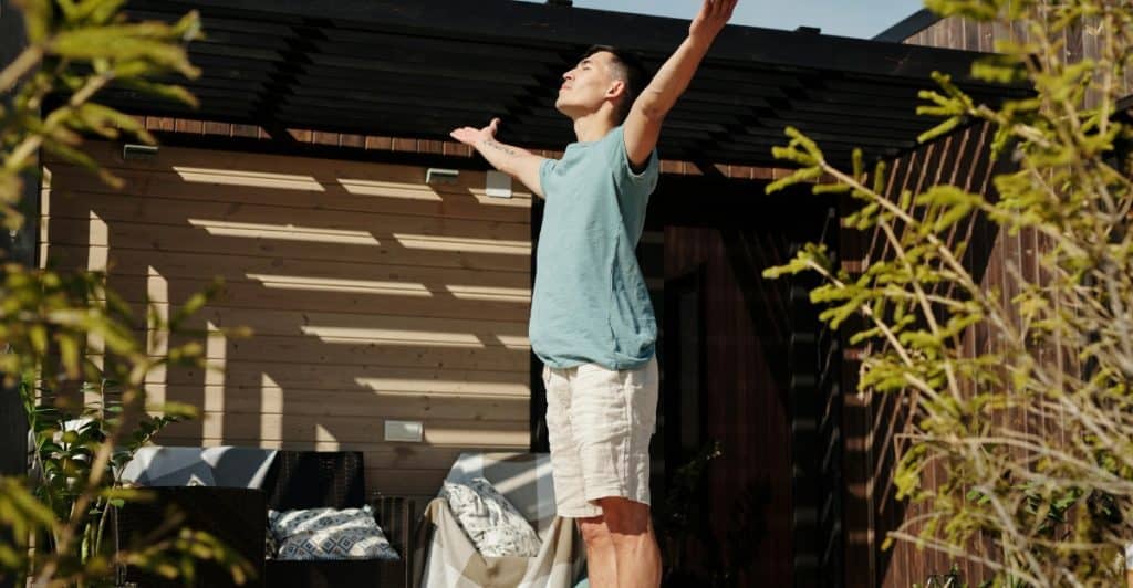 A man standing outdoors with his arms raised, eyes closed, seemingly stretching or practicing breathwork on a rug.