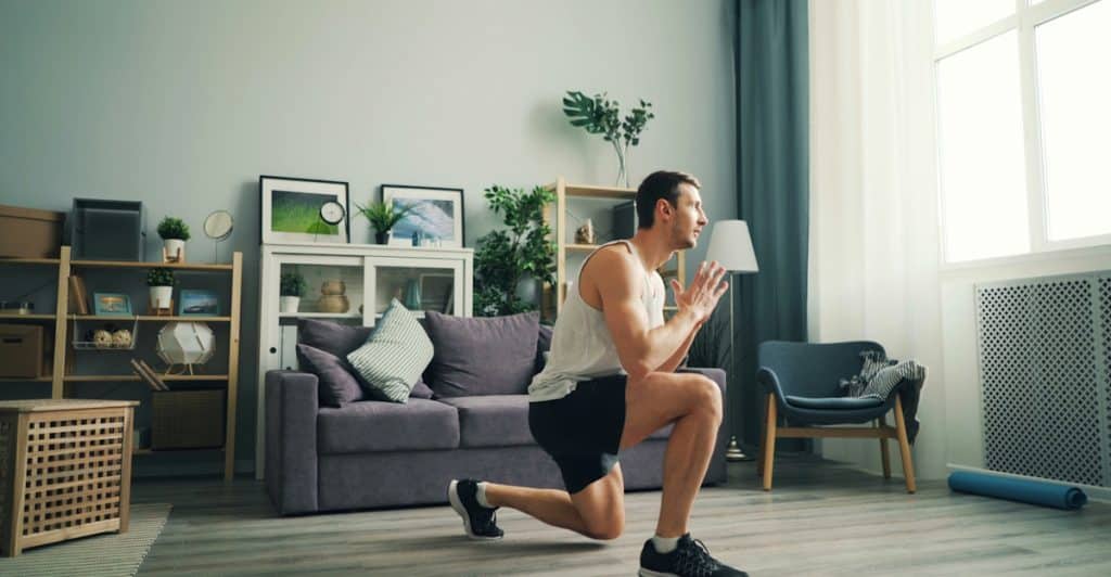 A man in a white tank top and black shorts doing lunges in a living room.