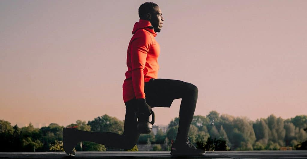 A man in a red hoodie and black pants performing a lunge while holding a kettlebell outdoors.