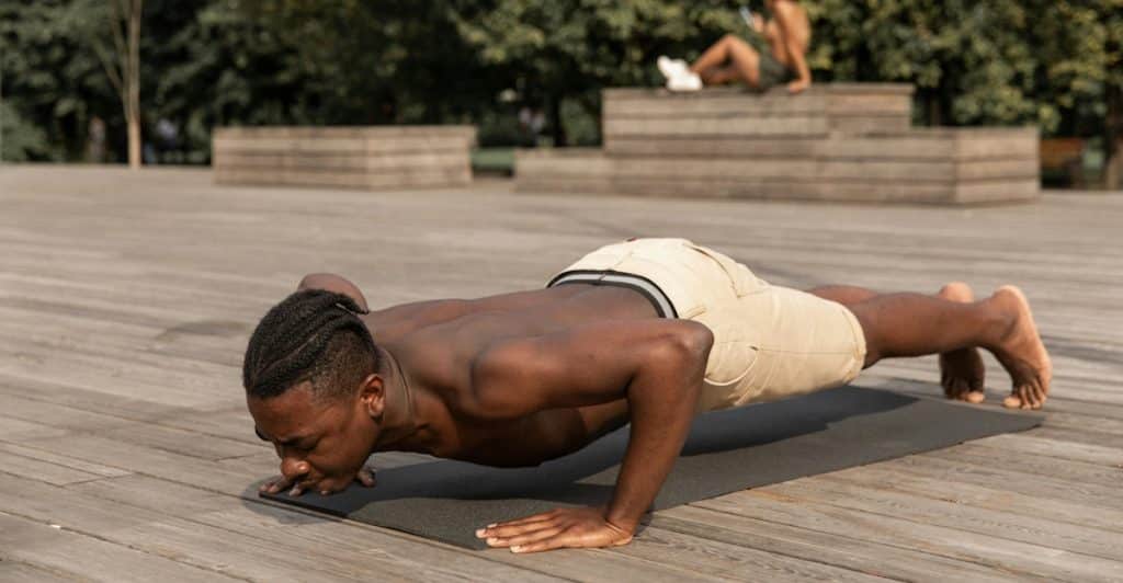 A shirtless man in beige shorts doing push-ups on a black yoga mat outdoors on a wooden deck.