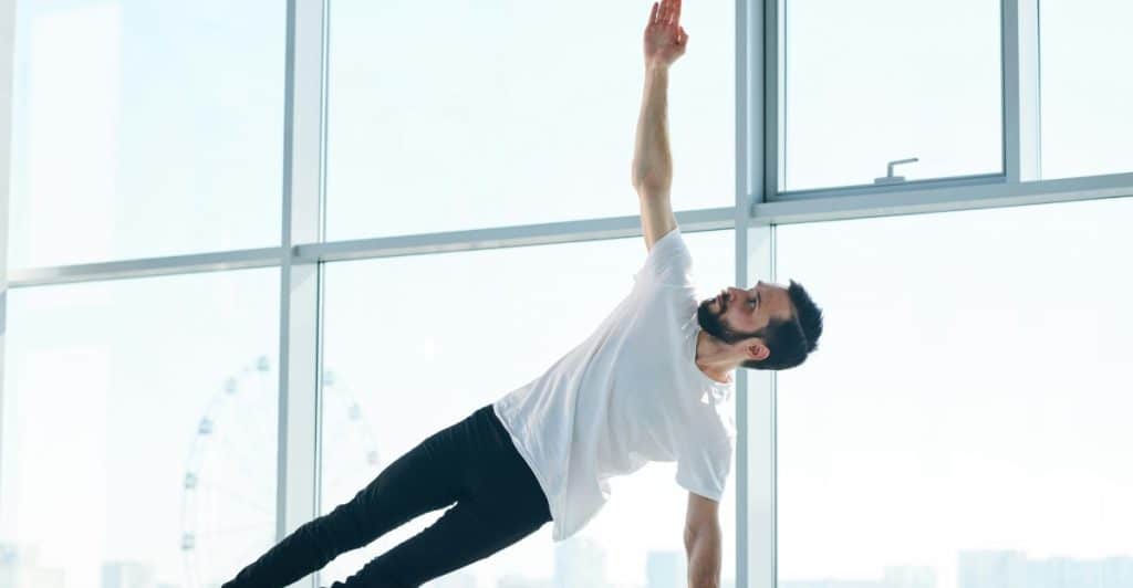 A bearded man in a white t-shirt and black pants is performing a side plank yoga pose on a black mat, with his right arm extended towards the ceiling, against a large window backdrop.