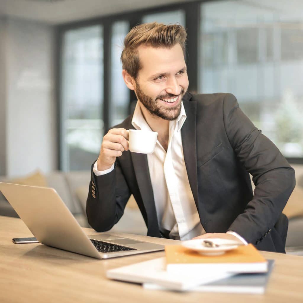 A smiling man in a suit jacket and open-collared shirt is seated at a light wooden desk, holding a white coffee cup to his mouth with his right hand, and looking towards the right while a silver laptop and some books are on the desk in front of him.