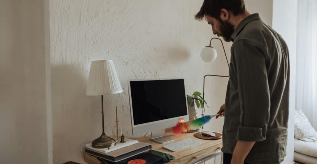 A man dusting a computer monitor and desk with a colorful duster
