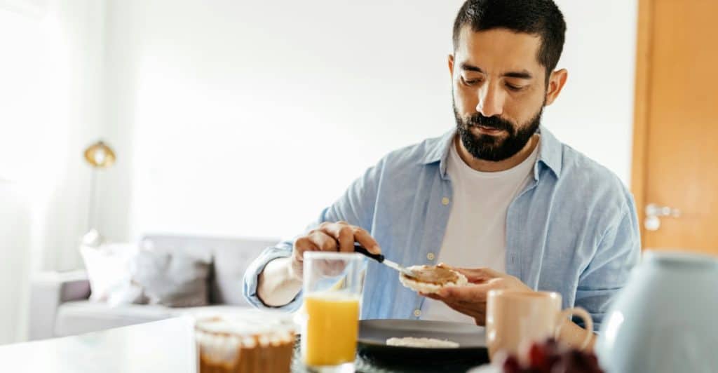 A man with a beard spreading something on toast while sitting at a kitchen counter with orange juice and fruit
