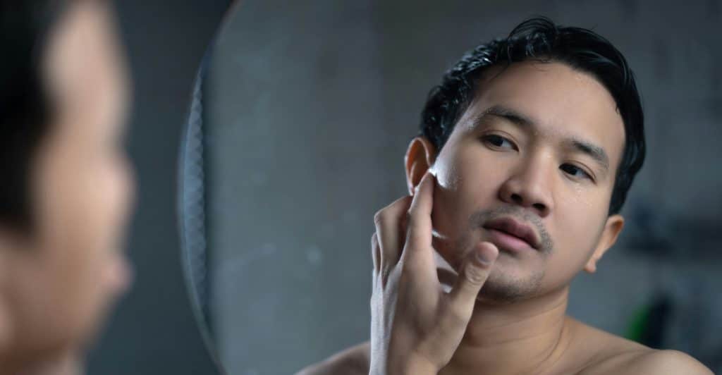 A shirtless man who appears to be of East Asian descent looking at his reflection in a round mirror while touching his cheek.