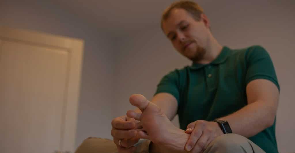 A man sitting down and carefully examining his foot with his hands.