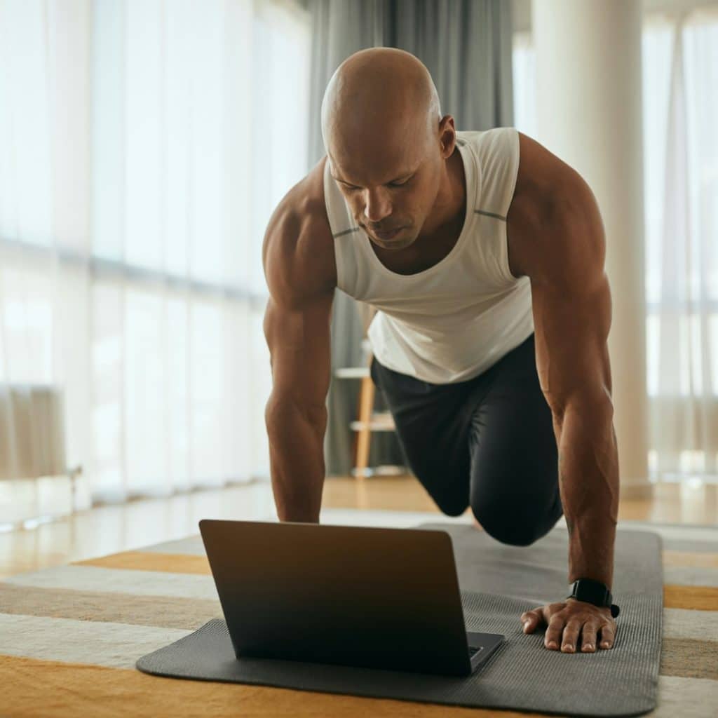 A man with dark hair and a beard is exercising outdoors, wearing a smartwatch on his left wrist, with his eyes closed and his head tilted back.