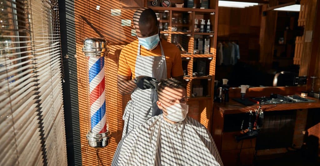 A barber wearing a mask and an apron is cutting a man's hair in a barbershop, with sunlight streaming through blinds and creating striped shadows.