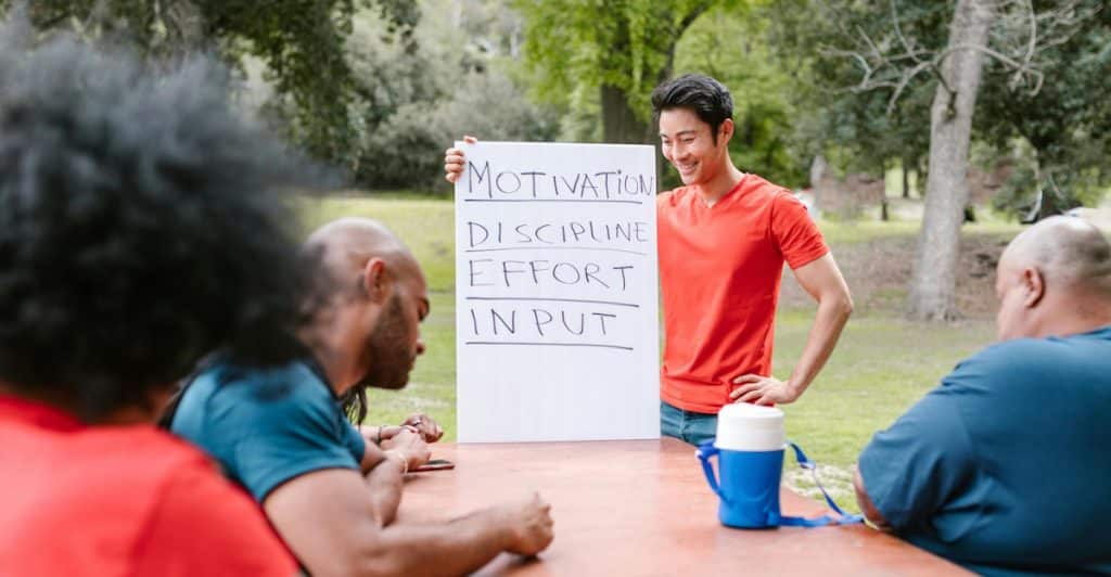 A man standing in front of a table with a white board that has "MOTIVATION," "DISCIPLINE," "EFFORT," and "INPUT" written on it, addressing a group of people sitting at the table.
