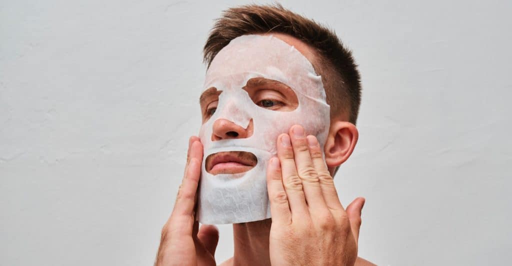 A man with short hair wearing a white tank top has a white sheet mask on his face and is touching it with both hands.