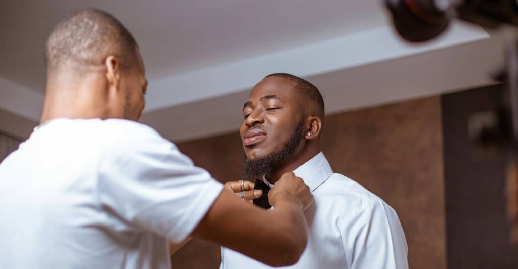One man in a white t-shirt is helping another man in a white dress shirt adjust his bow tie, with the second man's eyes closed and a camera visible on the right.