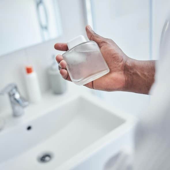 A man holding a fragrance bottle in a bathroom with a sink and mirror in the background.