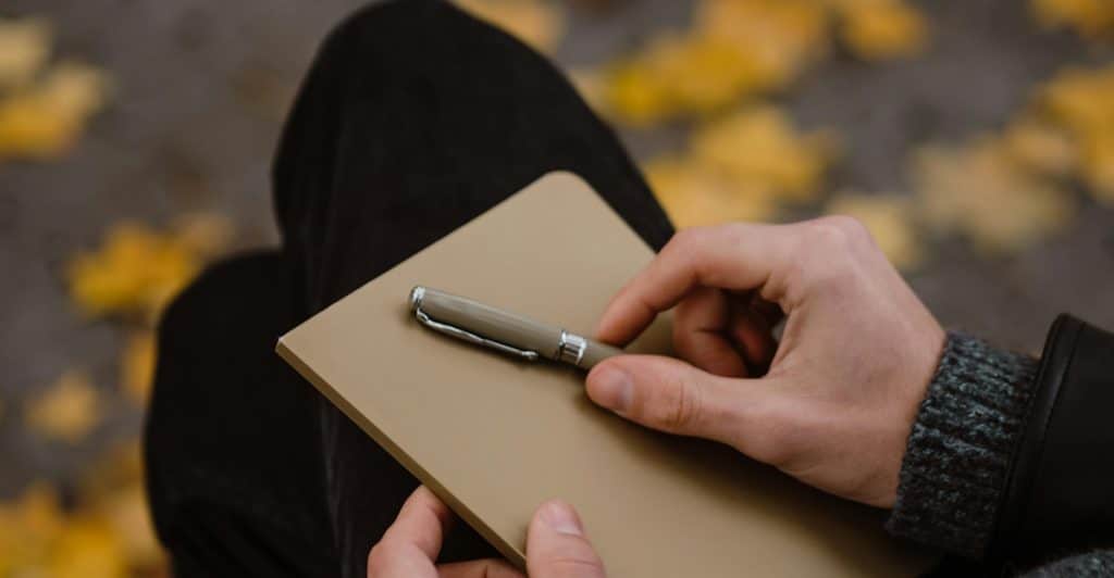 A man sitting outdoors, holding a notebook and pen, with blurred leaves in the background.