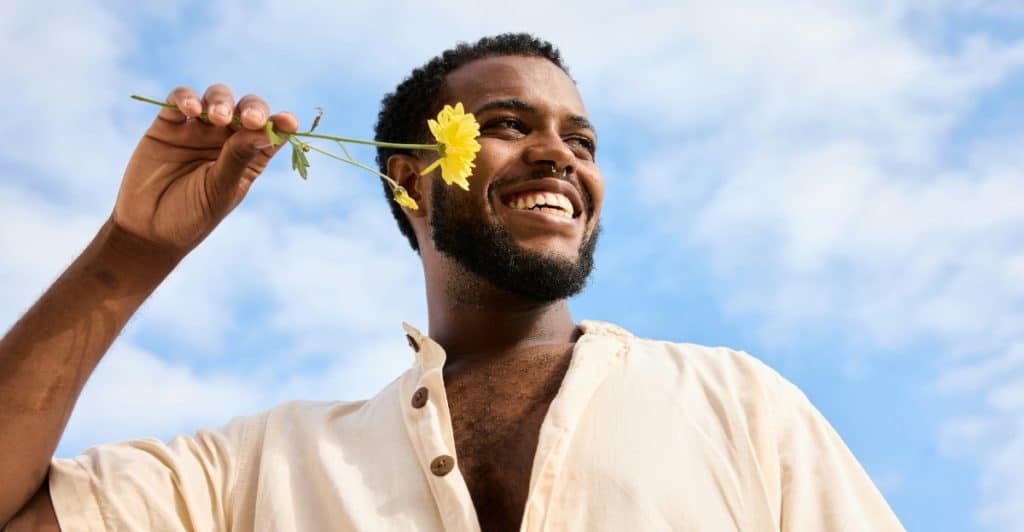 A man with a beard smiling and holding a yellow flower near his ear against a blue sky with white clouds.