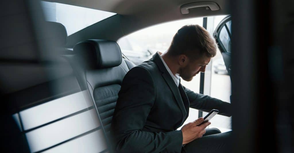 A man in a suit sitting in the backseat of a car, holding a smartphone and reaching toward the open door.