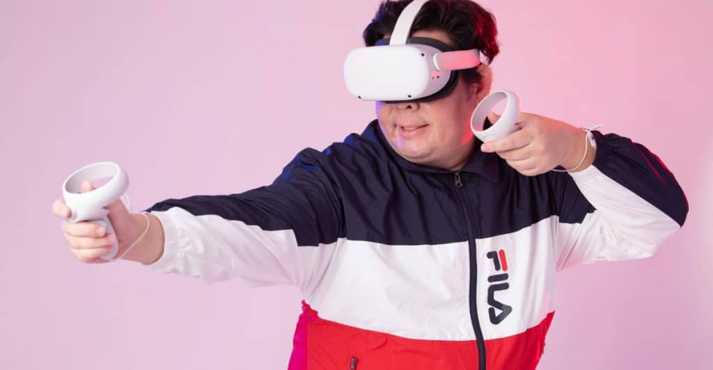 A man wearing a white VR headset and holding two white controllers, wearing a red, white, and blue Fila jacket against a pink background.