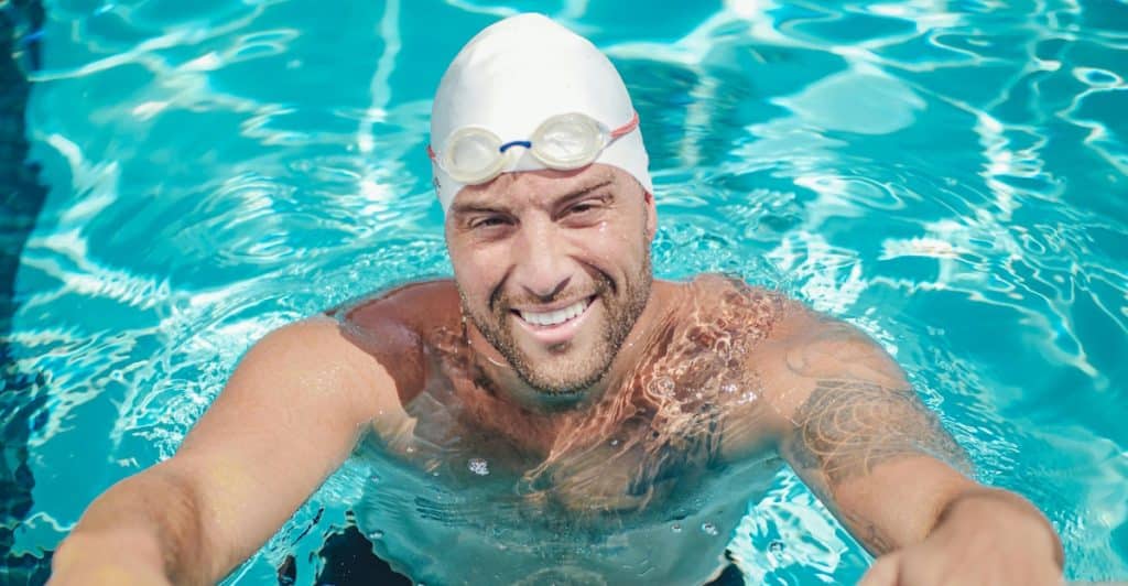 A smiling man wearing a swim cap and goggles in a swimming pool, holding onto the edge.