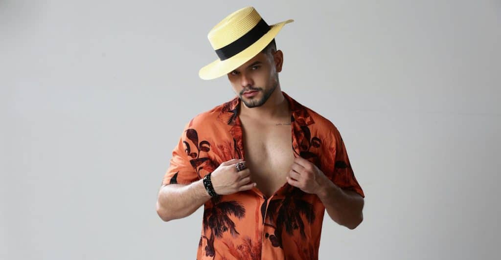 A man with a beard and a straw hat is looking at the camera, wearing an unbuttoned orange shirt with a deep V-neck.