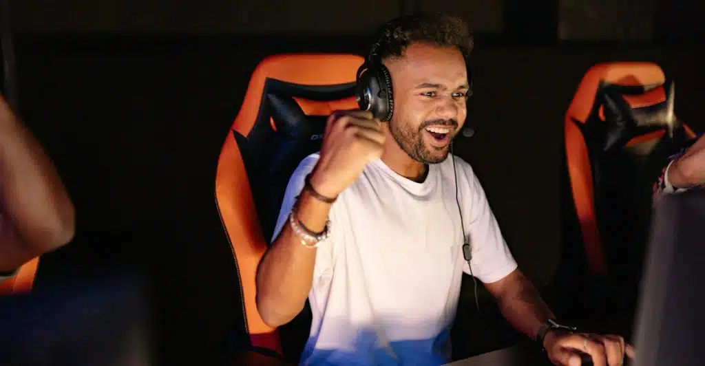 A man with headphones is sitting in an orange and black gaming chair, smiling while looking at a monitor, with another person partially visible next to him.