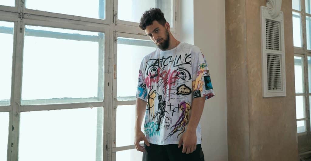 A man with a beard and curly hair is standing in front of large windows, wearing an oversized white graphic t-shirt and dark pants.