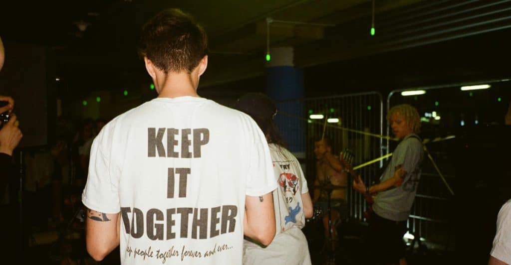 The back of a man wearing a white t-shirt with "KEEP IT TOGETHER" printed in large black letters is visible in a dimly lit, crowded indoor space.