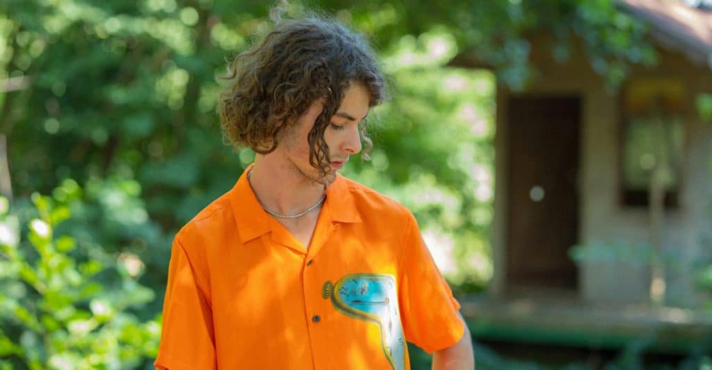 A man with curly hair is standing outdoors in front of greenery and a building, wearing a bright orange shirt with a light blue design on it.